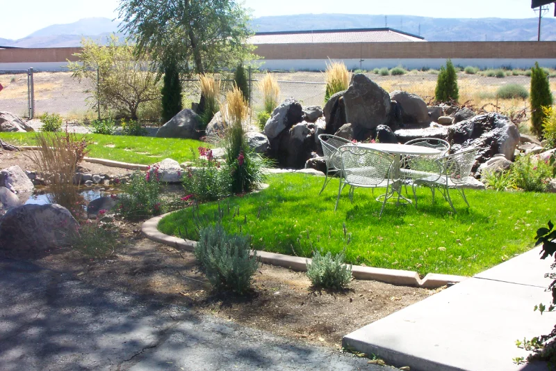 Backyard landscape with white patio furniture, boulders, pond, and bushes.