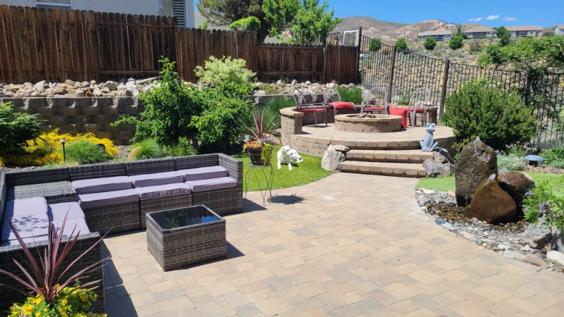 backyard landscape with wooden fence, stone patio, furniture, and a stone firepit with bushes and plants