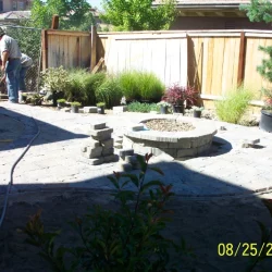 backyard patio area with a stone firepit, wooden dence, and men working in the background